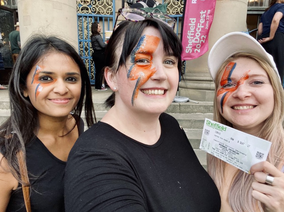 Bav, Kirsty, and Anna smile at the camera. Their faces are painted with glitter to recreate one of Bowie's iconic looks.
